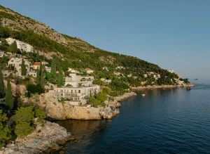 A modern white cliffside hotel built into rocky terraces overlooking the deep blue Adriatic Sea, with people sunbathing on the rocks below and green hills rising behind.