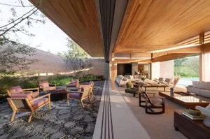 Modern covered outdoor living area featuring a stone-paved courtyard with fire pit, wooden lounge chairs with pink cushions, and a spacious open-plan interior lounge with warm wood ceilings and neutral furnishings.