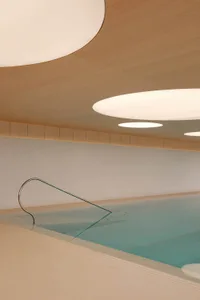 Modern indoor swimming pool with light blue water, stainless steel handrail, concrete walls and a curved wooden ceiling featuring large circular light fixtures.