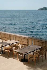 Outdoor dining tables and woven rattan chairs arranged on a stone terrace overlooking the calm blue sea with a distant boat and green island.