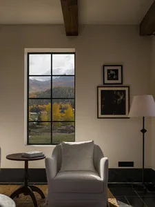 Cozy white armchair beside a large black-framed window overlooking autumn mountains and forest, with dark wood ceiling beams and black-and-white artwork on the wall.