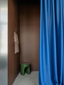 Modern minimalist entryway closet with terracotta plaster walls, a vibrant royal blue curtain, a glossy green sculptural stool, and a hanging beige t-shirt.