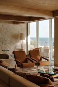 Modern coastal living room featuring a large travertine stone wall, two burnt orange lounge chairs, neutral sofa, glass coffee table, and panoramic ocean view through floor-to-ceiling windows.