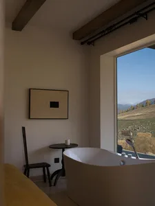 Modern minimalist bathroom featuring a white freestanding bathtub, dark wooden chair, small black side table, abstract artwork, and large window overlooking golden hills and mountains under a clear blue sky.