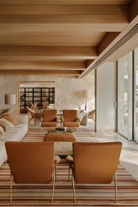 Modern living room featuring terracotta upholstered armchairs, a striped area rug, exposed wooden ceiling beams, and a large natural stone feature wall with built-in bookshelves in the background.
