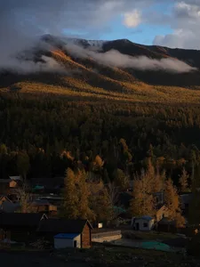 一座擁有許多木屋的偏遠山村坐落在廣闊森林山脈的山腳下，山林在金秋光線下閃耀，天空布滿戲劇性的雲層。
