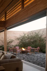 Modern outdoor courtyard with teak wood ceiling, irregular flagstone paving, a round metal fire pit with flames, and six wooden armchairs with blush pink cushions arranged around it.