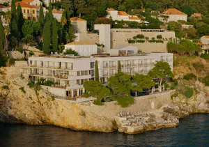 Modern white luxury hotel built into a rocky cliffside with multiple terraces, stone retaining walls, and direct access to the sea below.
