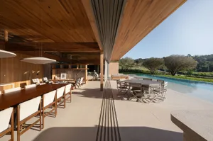 Modern covered patio featuring a long solid teak dining table, round outdoor dining set, infinity pool, and expansive wooden ceiling overlooking a green landscape.