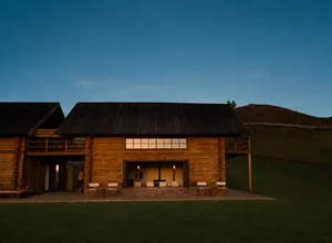 黃昏時分的大型雙層原木小屋外觀，室內燈光溫暖，外有休憩座椅區，背景為山景。
