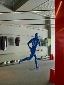 Interior view of a modern athletic apparel store featuring a glossy blue running mannequin, minimalist floating clothing racks with red accents, and linear LED lighting under an ornate ceiling.