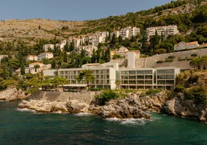 A contemporary seaside hotel built directly on a rocky coastline with turquoise water in the foreground and a hillside village rising behind it under a clear blue sky.