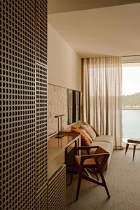 Modern coastal living room featuring a walnut console, wooden armchair, beige sofa with orange cushions, perforated wooden screen, and large window overlooking the sea.