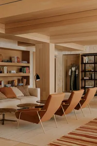 Modern minimalist living room featuring light oak wood paneling, built-in bookshelves, three cognac leather lounge chairs with brass legs, a cream sofa with terracotta cushions, and a striped area rug.