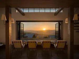 Interior view of a modern rustic sunroom with four wooden lounge chairs facing an open vista of mountains at sunrise, framed by heavy timber beams and large glass doors.