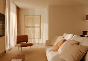 Minimalist living room featuring a cream boucle sofa with terracotta and beige cushions, a burnt orange leather accent chair with brass legs, two abstract watercolor artworks in wooden frames, sheer linen curtains, and warm natural sunlight creating soft shadows on beige walls.