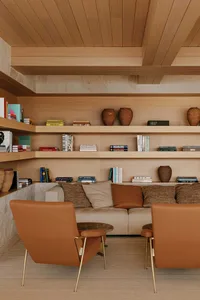 Modern living room featuring light oak built-in bookshelves, a beige sofa with terracotta and textured cushions, two burnt-orange leather armchairs with brass legs, and decorative pottery.