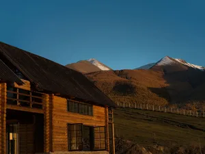 A rustic log cabin with dark wooden roof and large windows, bathed in golden sunset light, with snow-capped mountains and autumn-colored hills in the background.