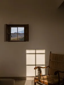 A wooden armchair with woven rattan seat and backrest placed beside a small open window revealing a scenic mountain landscape with autumn foliage and snow-capped peaks.
