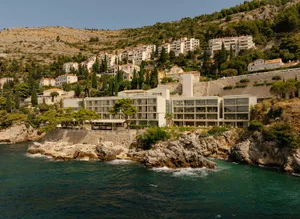 A contemporary white hotel with extensive glass windows built directly on rocky cliffs above the deep blue sea, with a hillside town in the background under a clear sky.