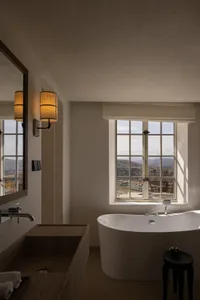 Modern bathroom interior featuring a white freestanding bathtub, wooden vanity with vessel sink, wall sconces, and large window overlooking mountains.