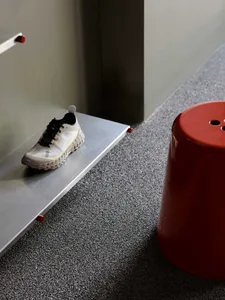 White and black athletic shoe resting on a stainless steel floating shelf above dark grey speckled carpet, with a bright red cylindrical stool beside it.