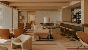 Spacious open-plan living room with travertine stone walls, terracotta leather armchairs, cream bouclé sofa, wooden ceiling beams, and a marble bar counter with leather stools.