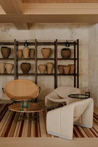 Modern wabi-sabi living room with two cream bouclé armchairs, a large round rattan peacock chair, marble coffee table, and a large dark wood shelving unit displaying numerous terracotta and earthenware pots against a textured stone wall.