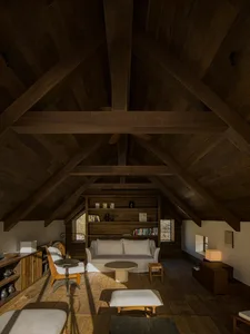 A spacious loft living room with high cathedral ceiling featuring massive reclaimed wood beams, a white sofa against a built-in wooden bookshelf, stone coffee table, and wooden flooring bathed in natural light.