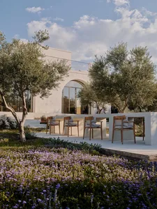 Modern Mediterranean villa terrace with wooden dining chairs, white limestone walls, olive trees, and a foreground of purple wildflowers under a bright blue sky.