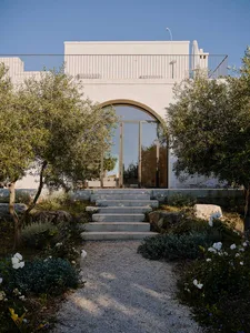 Front view of a modern white stucco villa with a large arched glass door, accessed by wide stone steps and surrounded by olive trees and Mediterranean garden plants under a clear blue sky.
