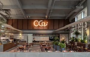 Modern industrial-style brunch and bistro interior with warm walnut wood paneling, concrete elements, upholstered seating, and numerous potted plants under bright natural and artificial lighting.
