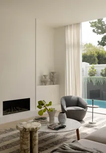 Minimalist living room featuring a marble fireplace, curved grey bouclé armchair, round marble coffee table, and large glass doors opening to a swimming pool and garden.