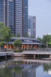 Modern curved waterfront pavilion with illuminated glass facade reflected in a pond at dusk, with high-rise buildings in the background.