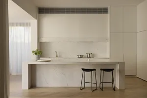 Modern minimalist kitchen featuring a large white marble island with two black wireframe stools, matching marble backsplash and counter, brass faucet, and soft natural light from a window with sheer curtains.