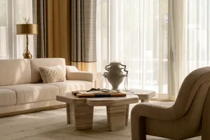 Bright minimalist living room featuring a cream boucle sofa, travertine coffee table, sculptural white vase, and sheer curtains with warm neutral tones.