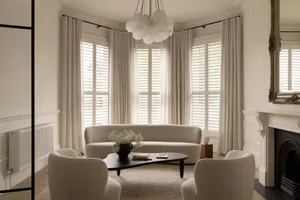 Bright and airy living room featuring a curved boucle sofa, two matching armchairs, a black organic coffee table, and a cloud-like chandelier in front of a large bay window with white shutters and curtains.