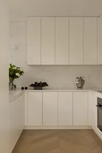 Modern minimalist kitchen featuring white flat-panel cabinetry, Calacatta marble backsplash and countertops, light herringbone wood flooring, and decorative objects including a flower vase and metallic sculpture.