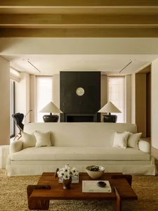 Modern minimalist living room featuring a large off-white sofa, dark walnut coffee table, black stone fireplace, and warm wooden ceiling beams.
