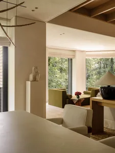 Modern living room interior featuring olive green sofas, light oak furniture, a stone sculpture on a pedestal, large windows overlooking a lush forest, and a black ceramic table lamp.