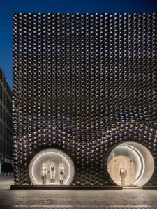 Exterior night view of a luxury retail building featuring a dramatic black perforated metal facade with two large illuminated circular entrances displaying mannequins.