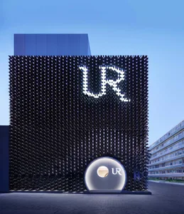 Night view of a modern building with a black perforated metal facade featuring a large illuminated UR logo and a circular entrance structure.
