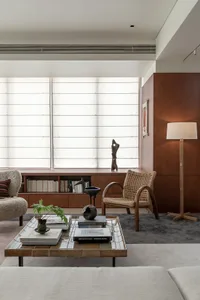 Modern living room featuring teak wood cabinetry, a woven rattan armchair, tiled coffee table, bouclé sofa, and large window with sheer blinds.