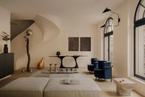 Modern organic living room featuring two large beige floor cushions, blue velvet armchairs, a black sculptural console table, a dramatic curved staircase, and a mix of natural materials including travertine and polished metal.