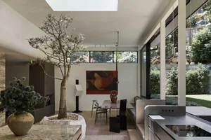 Modern open-plan dining room featuring a large indoor Plumeria tree planted in a stone planter, concrete architectural elements, a long marble dining table, built-in banquette seating, and large glass doors opening to a lush garden.