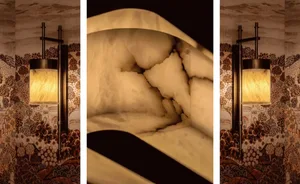 Close-up of a backlit onyx stone alcove with curved edges, flanked by two tall bronze wall sconces featuring cylindrical alabaster shades, set against luxurious hand-painted Chinoiserie wallpaper depicting trees, pagodas and landscapes.
