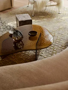 Close-up view of an irregular amber resin coffee table with textured surface, holding a crystal vase and small dish, placed on a beige patterned area rug in a modern living room.