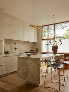 Modern minimalist kitchen featuring a large beige marble island, matching cabinetry, brass faucet and linear pendant light, with three leather and brass bar stools.