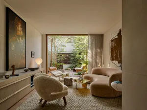 Modern living room featuring a curved boucle sofa, textured boucle armchair, stone coffee table, large abstract paintings, and an open view to a lush green courtyard garden.