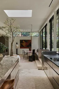 Modern minimalist dining room featuring a large indoor tree in a travertine planter, a pink marble dining table, built-in banquette, large skylight, and expansive glass walls overlooking the garden.